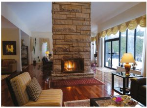 The residence’s comfortably appointed main reception rooms have three sitting areas (one just visible to left), a double-sided fireplace, a soaring stone chimney and a great view of the backyard. (Photo: Dyanne Wilson)